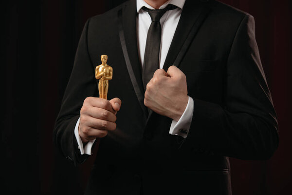 KYIV, UKRAINE - JANUARY 10, 2019: partial view of man in suit with clenched fist holding oscar award on dark background