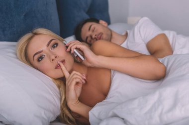 Smartphone ile konuşmak ve kocası yatakta, gizli kavramı uyku sırasında dudaklarında parmak ile kameraya bakıyor güzel genç kadın 