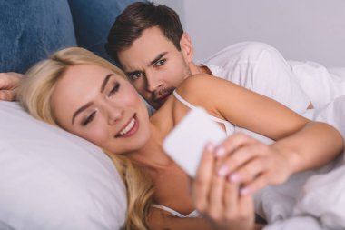 şüpheli genç adam gülümseyen karısı yatakta, güvensizlik kavram smartphone kullanarak gözatma 