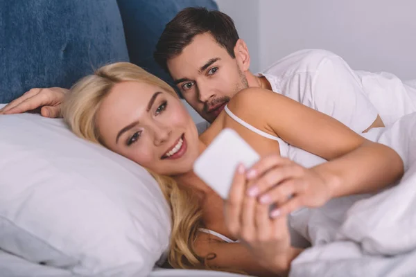 Jovem Espreitando Namorada Sorrindo Usando Smartphone Cama Conceito Desconfiança — Fotografia de Stock