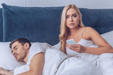 akıllı telefon tutarak ve kamera erkek yatakta, güvensizlik kavramı uyku sırasında bakarak mutsuz genç kadın
