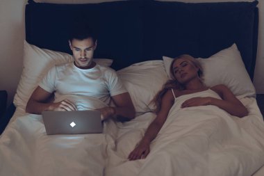 Genç adamın karısı yatakta gece uyku sırasında dizüstü kullanarak yüksek açılı görünüş