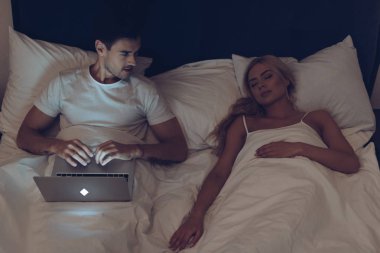 bir dizüstü bilgisayar kullanarak ve geceleri yatakta uyuyan eşi bakarak Genç adam yüksek açılı görünüş