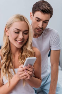 Yatak odasında smartphone kullanarak güzel gülümseyen karısı bakarak kıskanç genç adam 