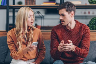 shocked young woman holding smartphone and looking at husband sitting on couch, jealousy concept clipart