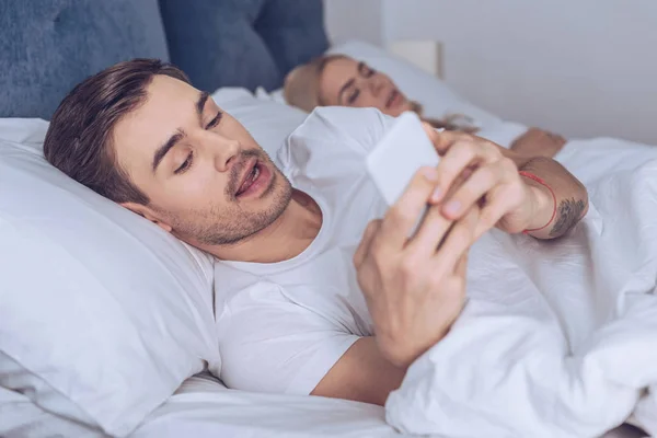 Suspicious Young Man Using Smartphone While Lying Sleeping Wife Bed — Stock Photo, Image
