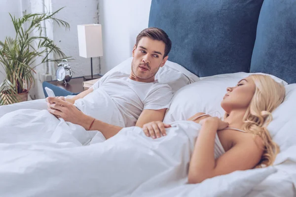 Homem Suspeito Deitado Cama Com Smartphone Olhando Para Namorada Dormindo — Fotografia de Stock