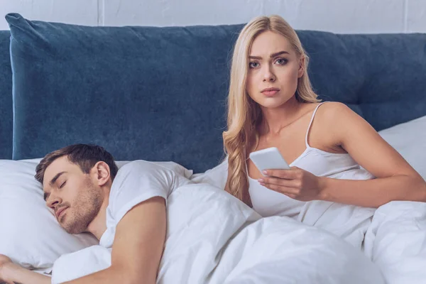 Unhappy Young Woman Holding Smartphone Looking Camera While Boyfriend Sleeping — Stock Photo, Image