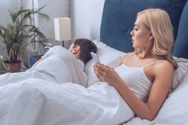 Jealous Young Woman Using Smartphone Looking Boyfriend Sleeping Bed Distrust — Stock Photo, Image