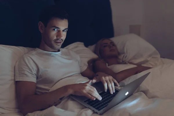 Joven Sospechoso Usando Ordenador Portátil Mirando Cámara Mientras Esposa Duerme — Foto de Stock