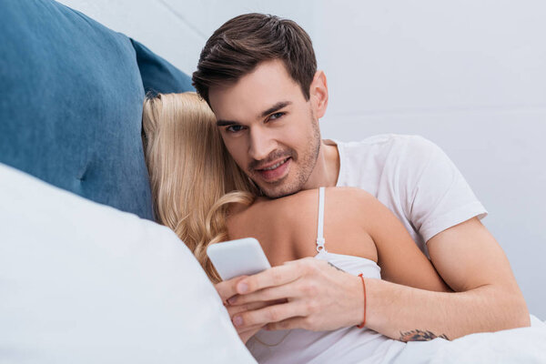 young man using smartphone and smiling at camera while lying with girlfriend in bed