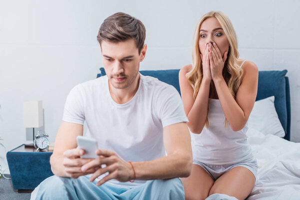 shocked young woman looking at boyfriend using smartphone on bed, distrust concept