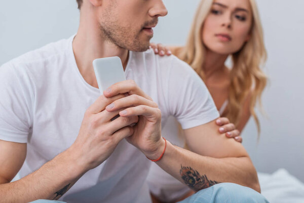 cropped shot of man using smartphone and girlfriend peeking behind in bedroom, distrust concept
