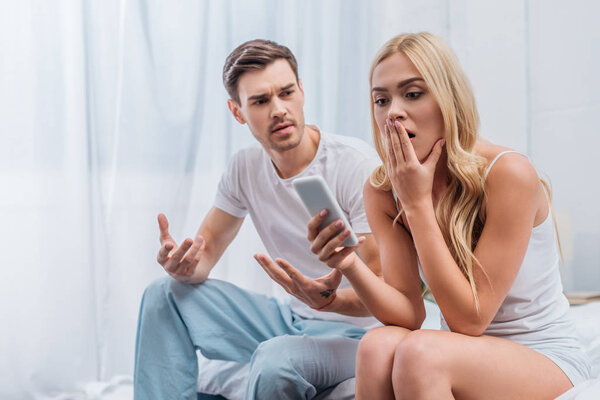 emotional young man gesturing with hands and looking at shocked girlfriend using smartphone on bed