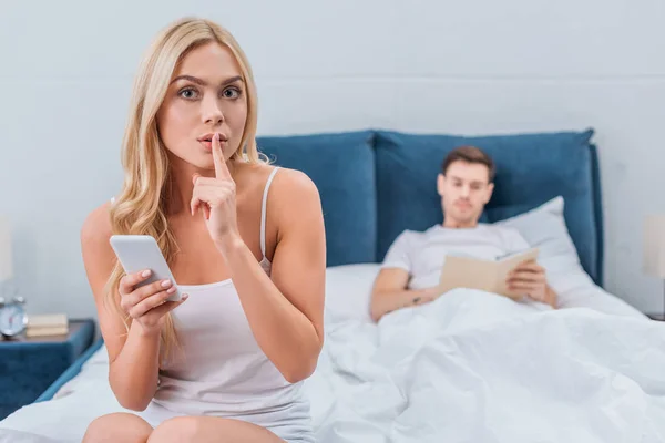 Young Woman Smartphone Gesturing Silence Looking Camera While Boyfriend Reading — Stock Photo, Image