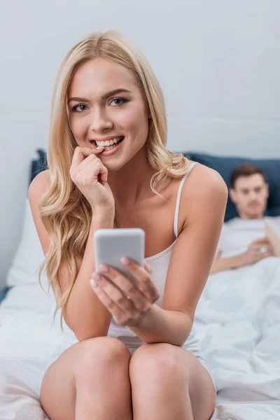 Jonge Vrouw Met Smartphone Nagel Bijten Glimlachend Camera Terwijl Echtgenoot — Stockfoto