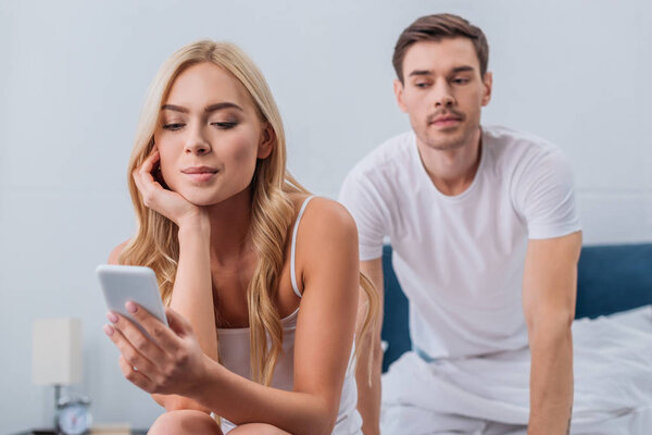 улыбающаяся молодая женщина с помощью смартфона и ревнивый мужчина подглядывает сзади на кровати
 