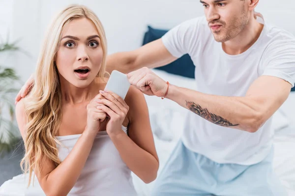 Jealous Man Grabbing Smartphone Scared Girlfriend Looking Camera Bedroom — Stock Photo, Image