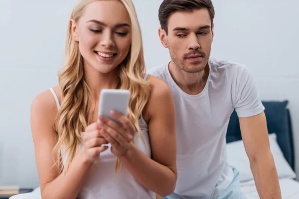 Jealous Man Looking Beautiful Smiling Wife Using Smartphone Bedroom Mistrust — Stock Photo, Image