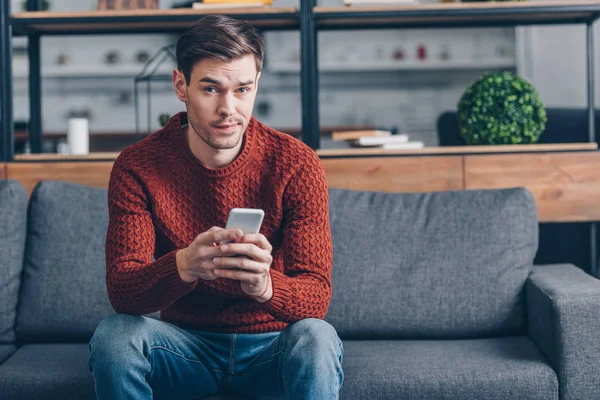 Suspicious Young Man Holding Smartphone Looking Camera While Sitting Couch — Stock Photo, Image