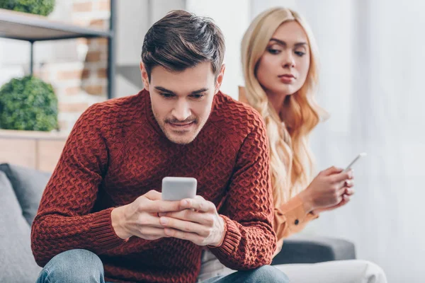 嫉妒的年轻女子与智能手机看微笑的男友在家里使用智能手机 关系问题的概念 — 图库照片