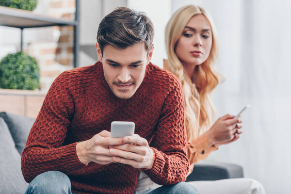 jealous young woman with smartphone looking at smiling boyfriend using smartphone at home, relationship problem concept