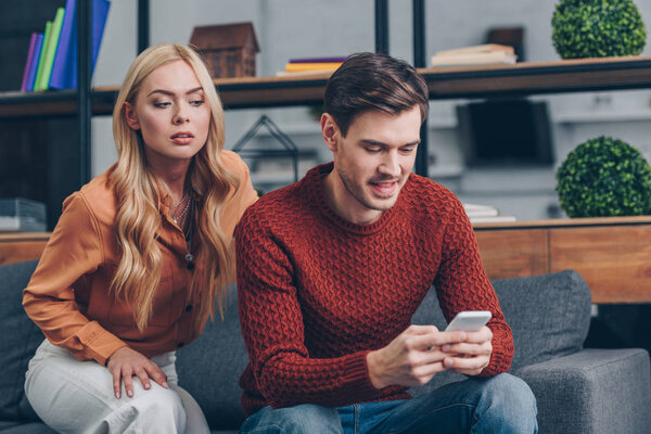 jealous young woman looking at smiling husband using smartphone on couch, distrust concept