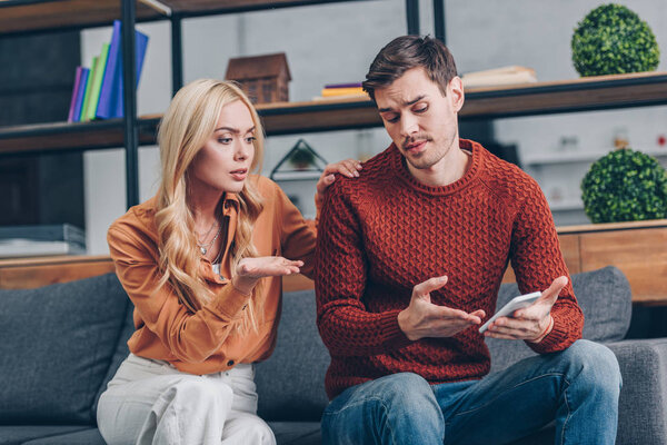 upset young couple sitting on couch and discussing smartphone, distrust concept