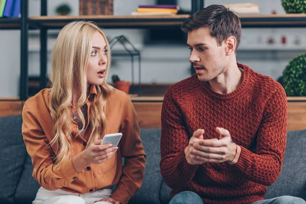 Shocked Young Woman Holding Smartphone Looking Husband Sitting Couch Jealousy — Stock Photo, Image