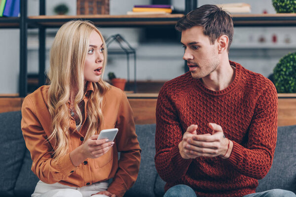 shocked young woman holding smartphone and looking at husband sitting on couch, jealousy concept