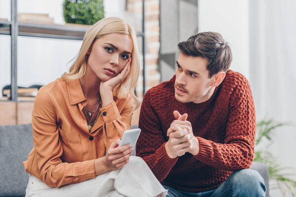 муж смотрит на несчастную молодую женщину, держащую смартфон и глядя на камеру дома, концепция проблемы отношений
