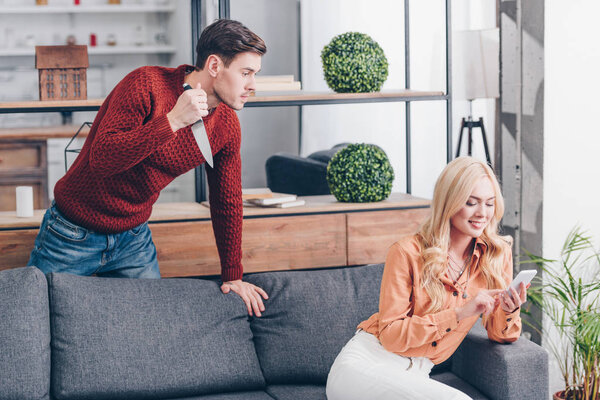 агрессивный мужчина с ножом глядя на улыбающуюся жену с помощью смартфона дома, концепция недоверия
 