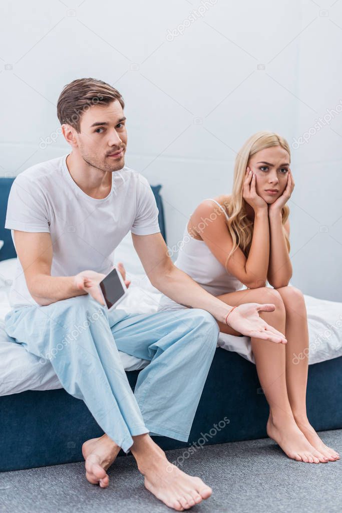 upset young woman sitting on bed while boyfriend holding smartphone and looking at camera, mistrust concept