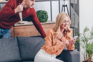 cropped shot of aggressive man with knife looking at smiling wife using smartphone on couch, jealousy concept  clipart