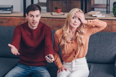 jealous man with smartphone and upset woman sitting on couch and looking at camera, distrust concept clipart
