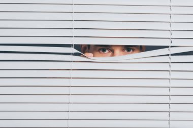 suspicious young man peeking and looking at camera through blinds, mistrust concept clipart