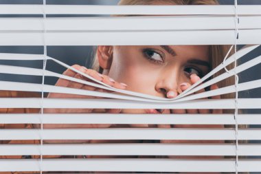 young woman looking away and peeking through blinds, mistrust concept  clipart