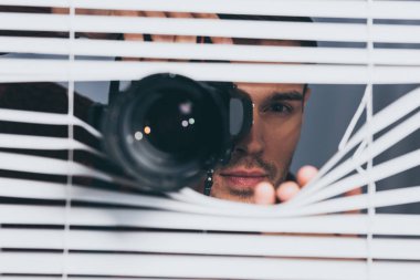 young man holding camera and looking at camera through blinds, mistrust concept clipart