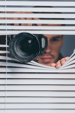 close-up view of young man holding camera and looking at camera through blinds clipart