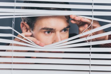 suspicious young man looking away through blinds, spying concept clipart