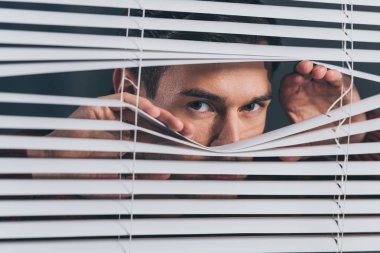 young man spying and looking at camera through blinds clipart