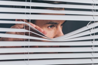 close-up view of young man spying and looking away through blinds clipart