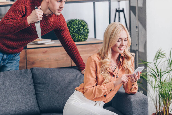 cropped shot of aggressive man with knife looking at smiling wife using smartphone on couch, jealousy concept 