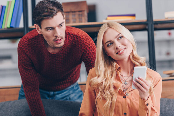 шокированный муж смотрит на красивую улыбающуюся жену с помощью смартфона дома, концепция ревности
 