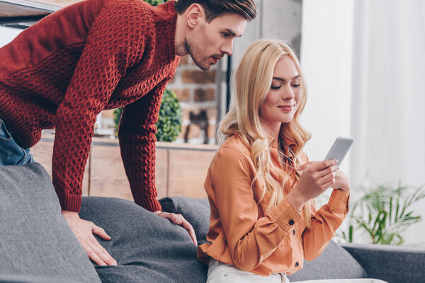 ревнивый молодой человек смотрит на улыбающуюся девушку, сидящую на диване и использующую смартфон, концепцию недоверия
