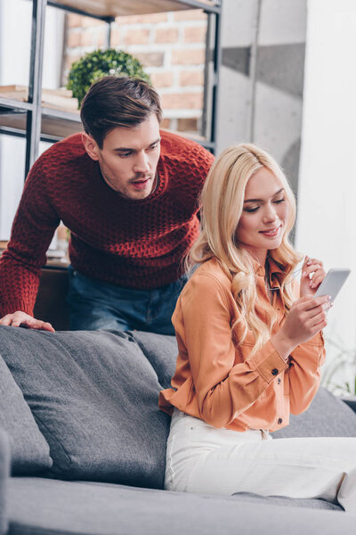 jealous emotional man looking at smiling young wife using smartphone at home, mistrust concept