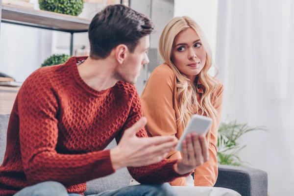 Joven Emocional Sosteniendo Teléfono Inteligente Mirando Novia Confundida Sentada Sofá —  Fotos de Stock