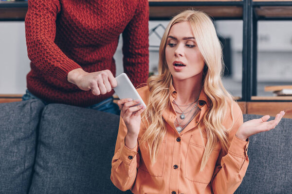 обрезанный снимок мужчины, берущего смартфон от эмоциональной подружки, сидящей на диване, концепция ревности
