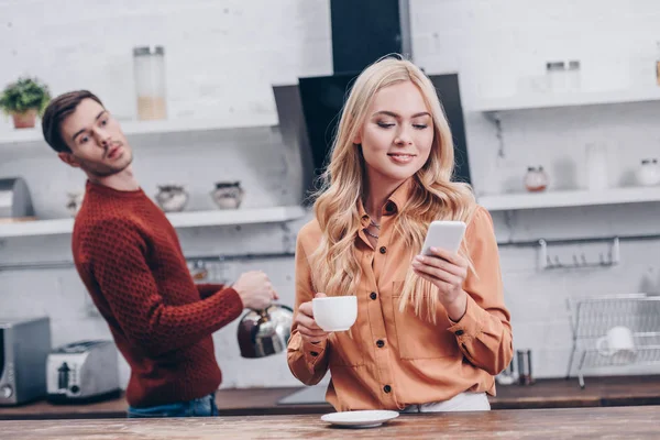 Joven Celoso Mirando Sonriente Novia Sosteniendo Taza Uso Teléfono Inteligente —  Fotos de Stock