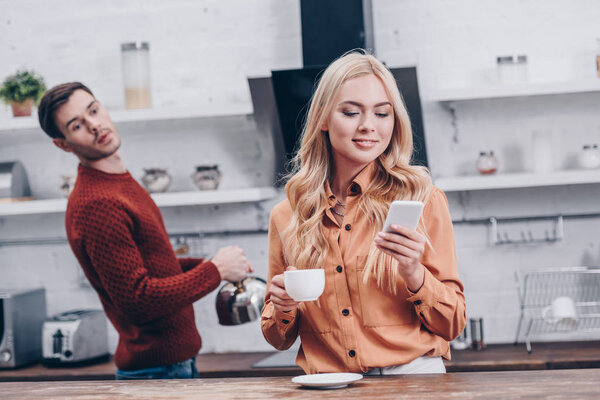jealous young man looking at smiling girlfriend holding cup and using smartphone in kitchen